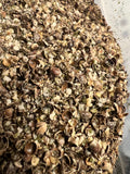 Angled close-up of a large quantity of raw organic crushed hemp seeds in a clear plastic container, showing cracked dark brown and tan seed shells with visible green hemp hearts and fine seed meal, with the container edge visible in the background
