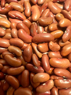 Macro close-up of bulk red kidney beans piled together, showing large plump elongated beans in deep red and auburn tones with smooth shiny seed coats, white hilum markings, and natural variation in size and curvature